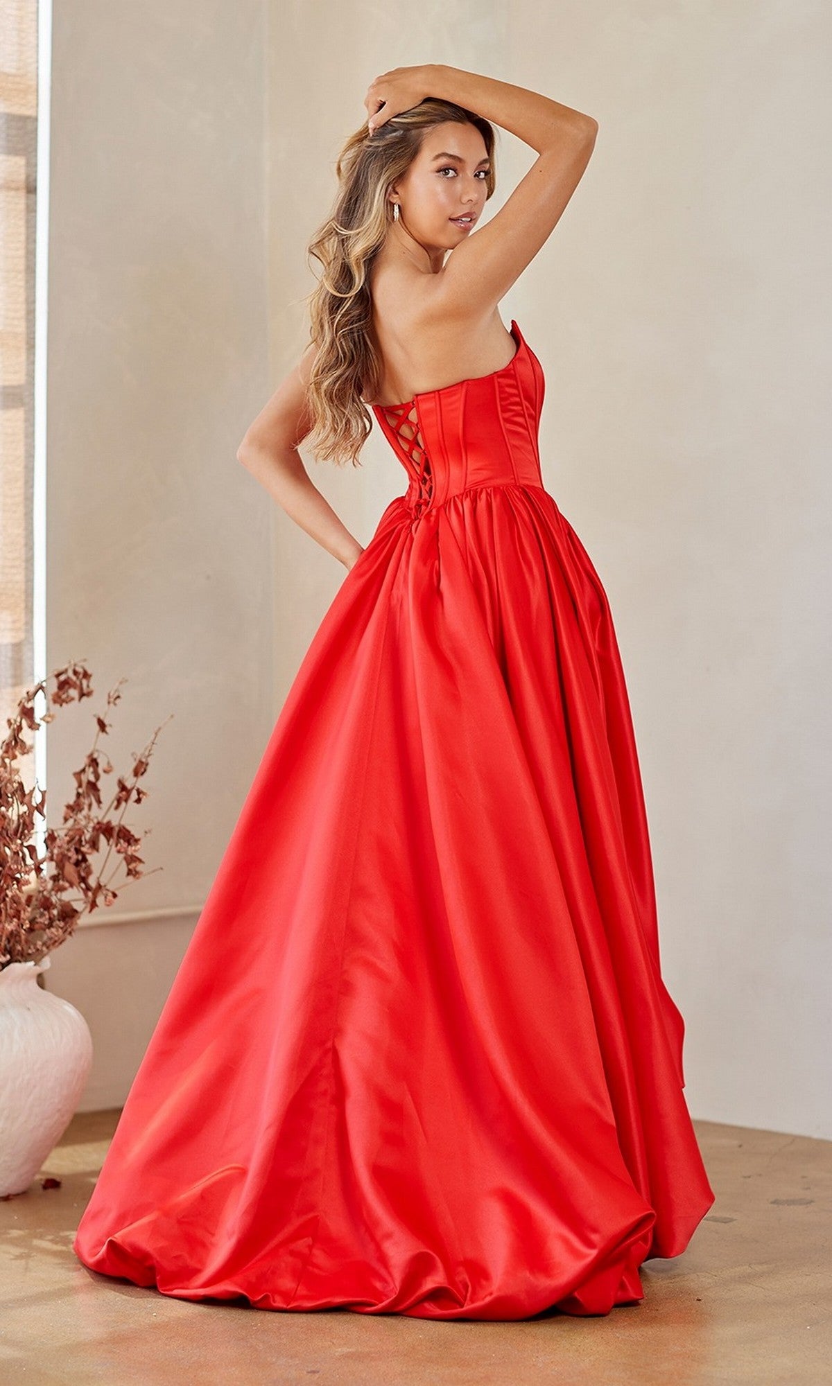 A woman wearing the Amelia Couture 2085 long prom dress, a strapless floor-length gown with a corset bodice, stands indoors looking over her shoulder with one hand raised to her head.